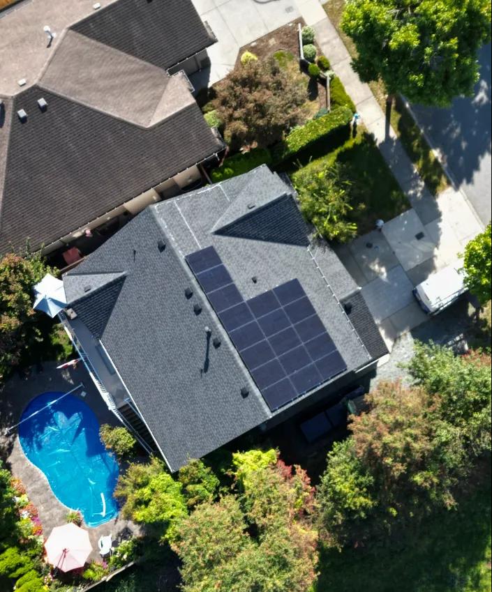 Aerial drone view of a modern residential home featuring a sleek rooftop solar panel installation next to a backyard swimming pool.