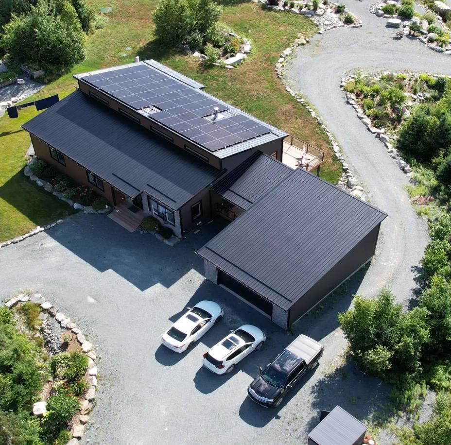 An aerial drone view of a modern residential home situated in a lush, wooded landscape, featuring a large black solar panel installation across its roof.